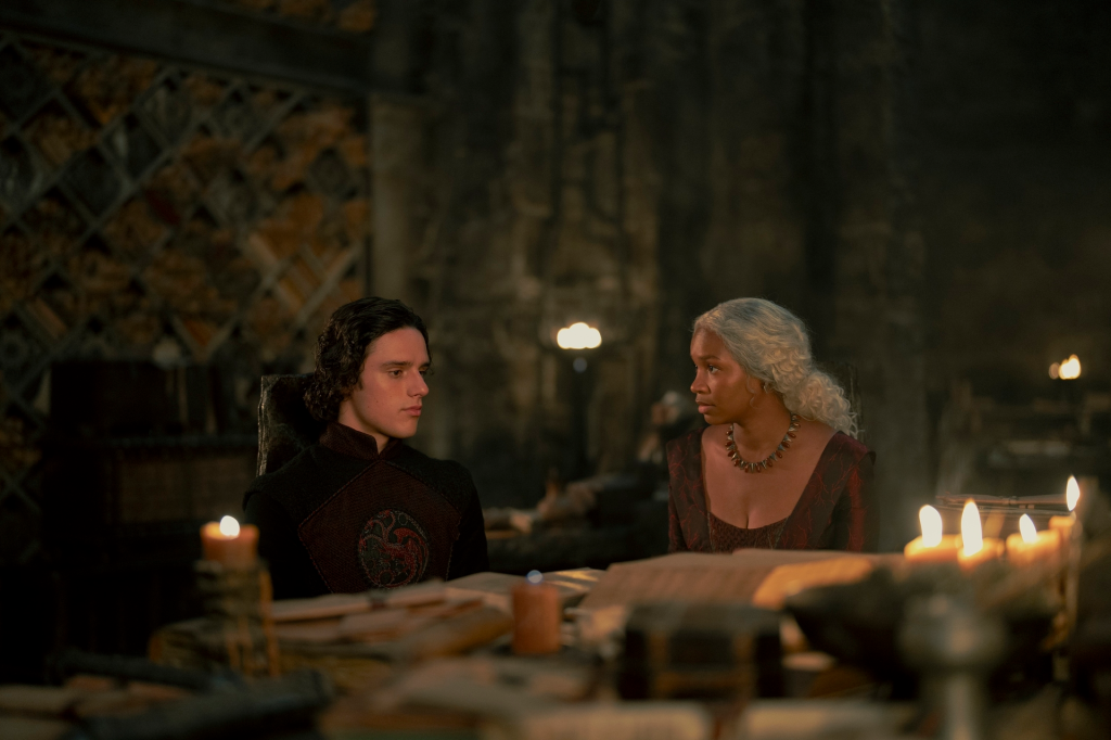 Harry Collett as Jacaerys Velaryon and an unnamed woman sitting at a table with books and candles in 