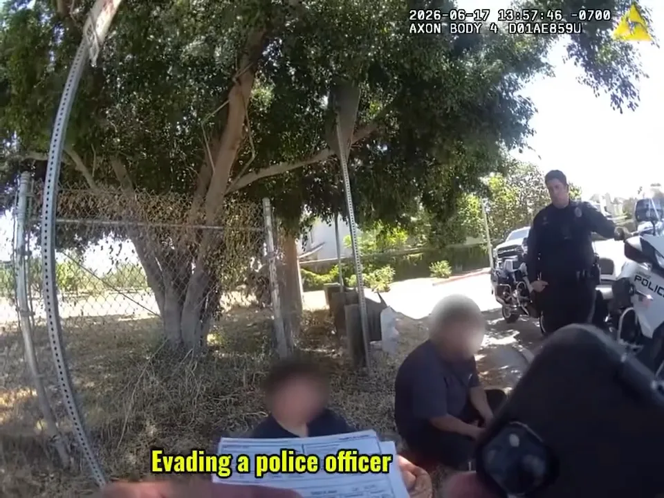 Footage from an officer's body camera showing a police officer next to his motorcycle while two individuals sit on the ground, with text 