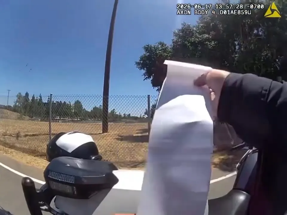 A police body camera recording shows a hand holding paperwork near a motorcycle, with a chain-link fence and trees in the background.