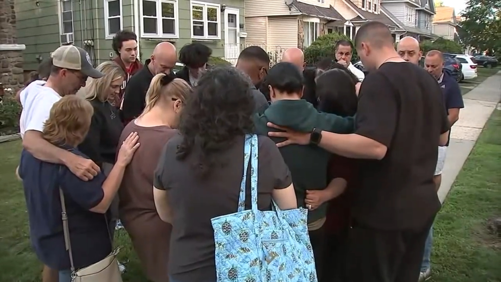 A group of people standing close together in a circle, some with their arms around each other, with houses and a street in the background.
