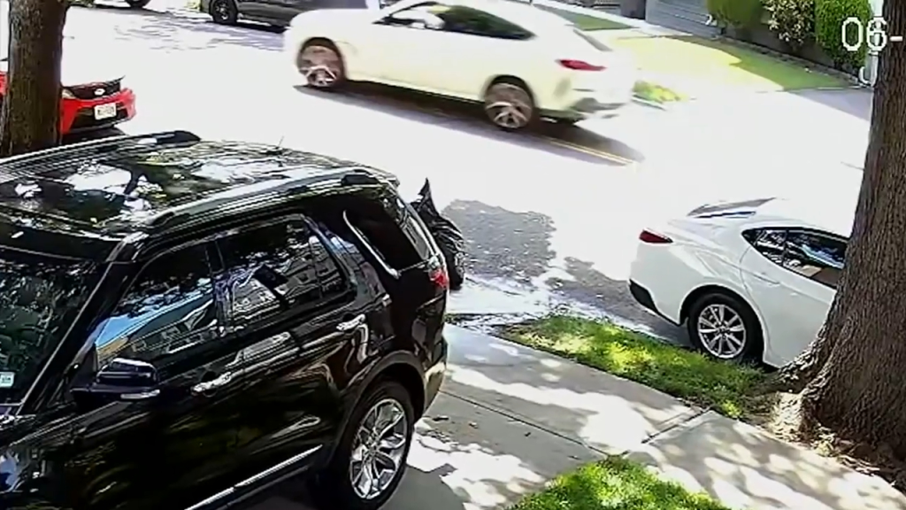 A white BMW speeding down a residential street, passing a parked black SUV and white car.