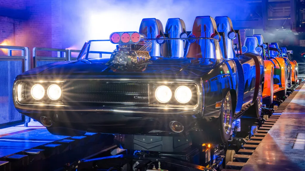 Roller coaster cars designed as classic muscle cars, including a black Dodge Charger, lined up on a track.
