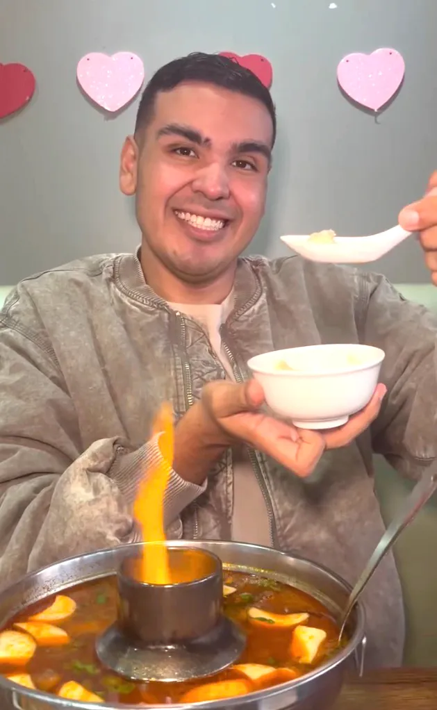 A man smiling while holding a spoon with food and a bowl of food next to a flaming hot pot.