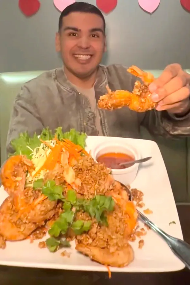 Man holding up a fried shrimp with a full plate of garlic butter fried shrimp and sauce.