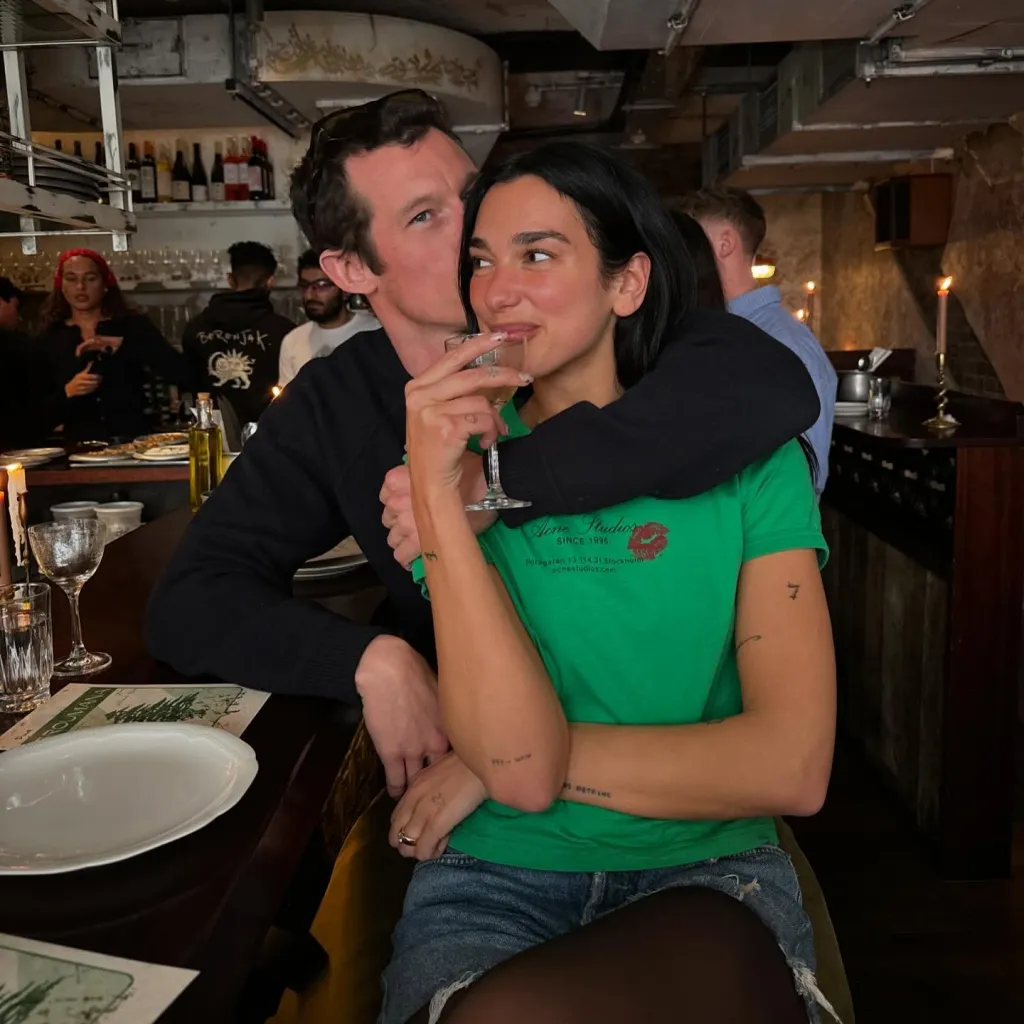 Callum Turner embracing Dua Lipa from behind at a restaurant.