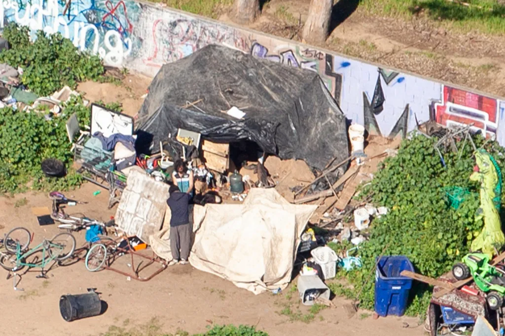 Overhead view of a homeless encampment with a large black tarp tent, surrounded by various discarded items, bicycles, and green bushes, against a graffiti-covered wall.