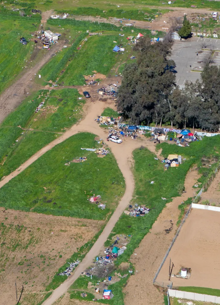 Aerial view of a homeless encampment with tents, tarps, vehicles, and trash scattered across green and brown fields.