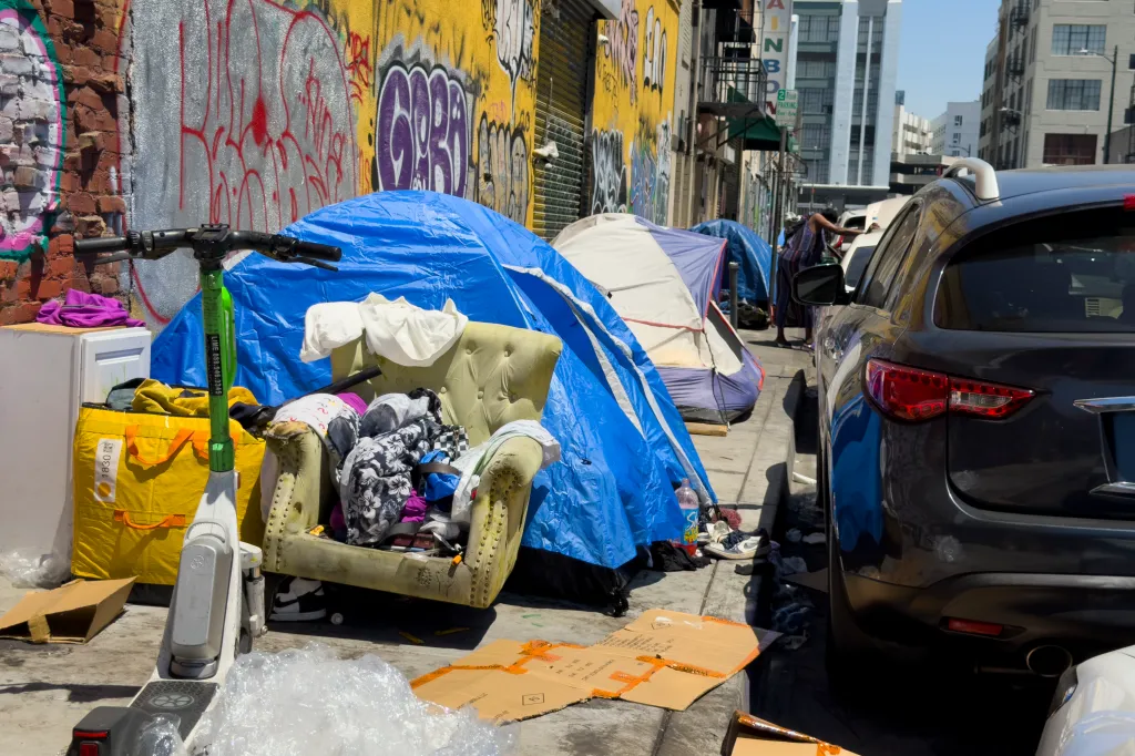 Tents, an armchair filled with belongings, and an electric scooter line a graffiti-covered sidewalk in Skid Row, Los Angeles.
