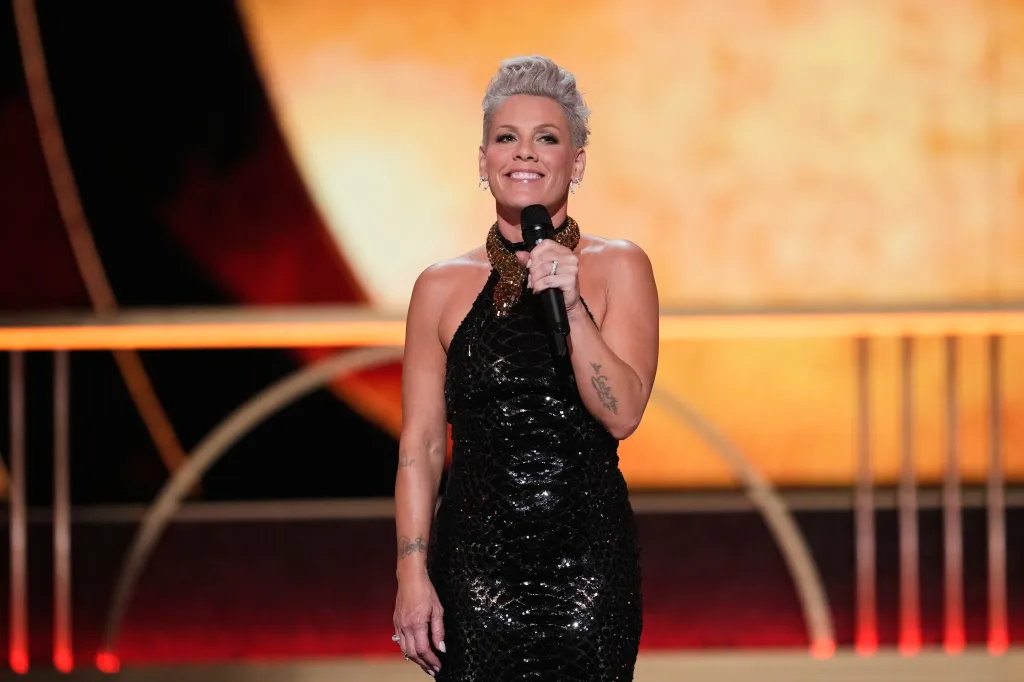 Pink, wearing a black sequined dress with a gold snake-like neckpiece, speaks into a microphone as host of the 79th Tony Awards.