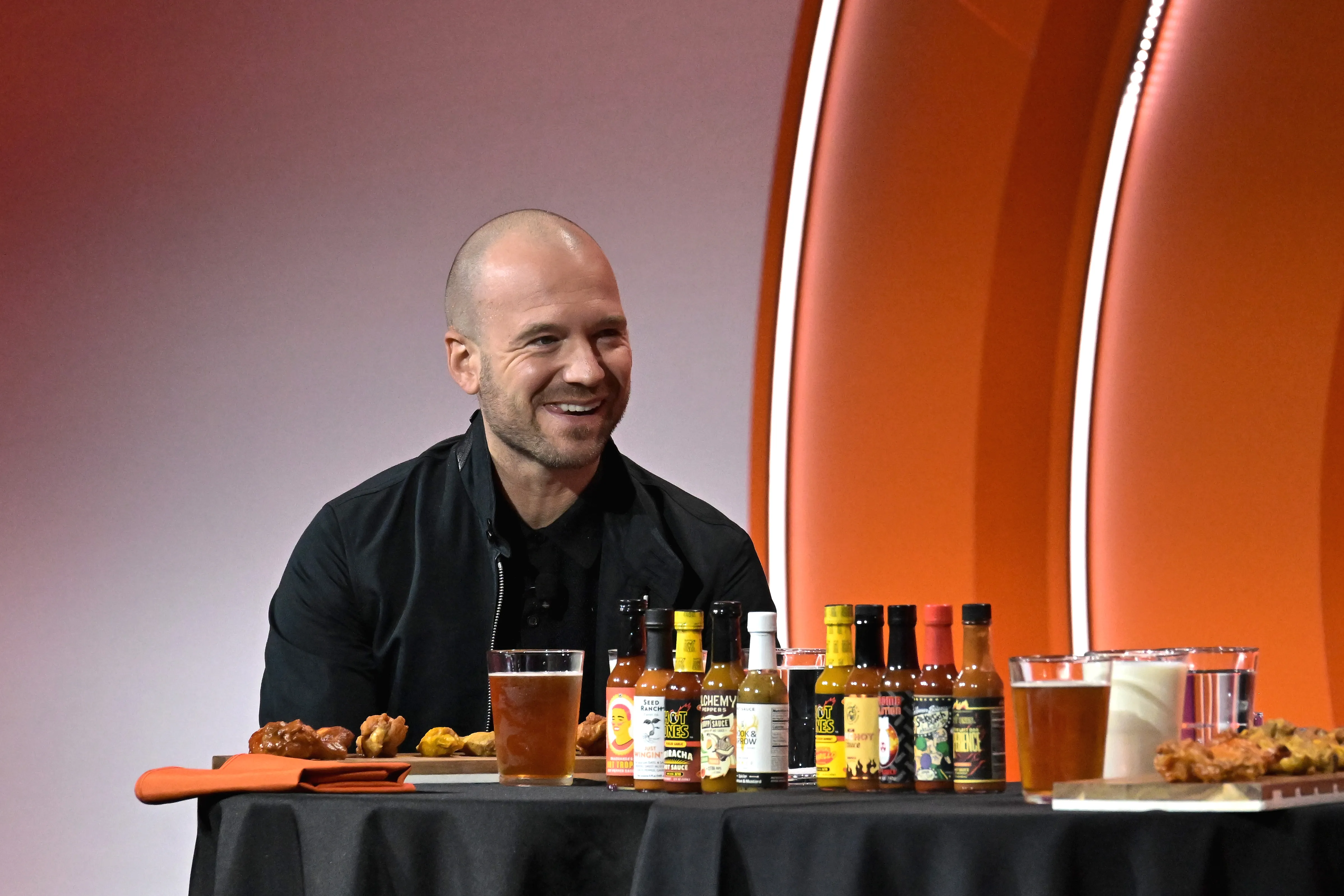 Sean Evans, host of Hot Ones, smiles while seated at a table with hot sauces, chicken wings, and drinks.