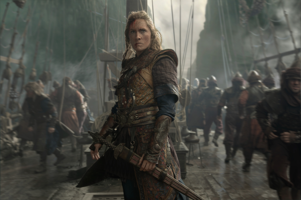 A bloodied woman in armor holds a sword on a ship as armed men stand behind her.