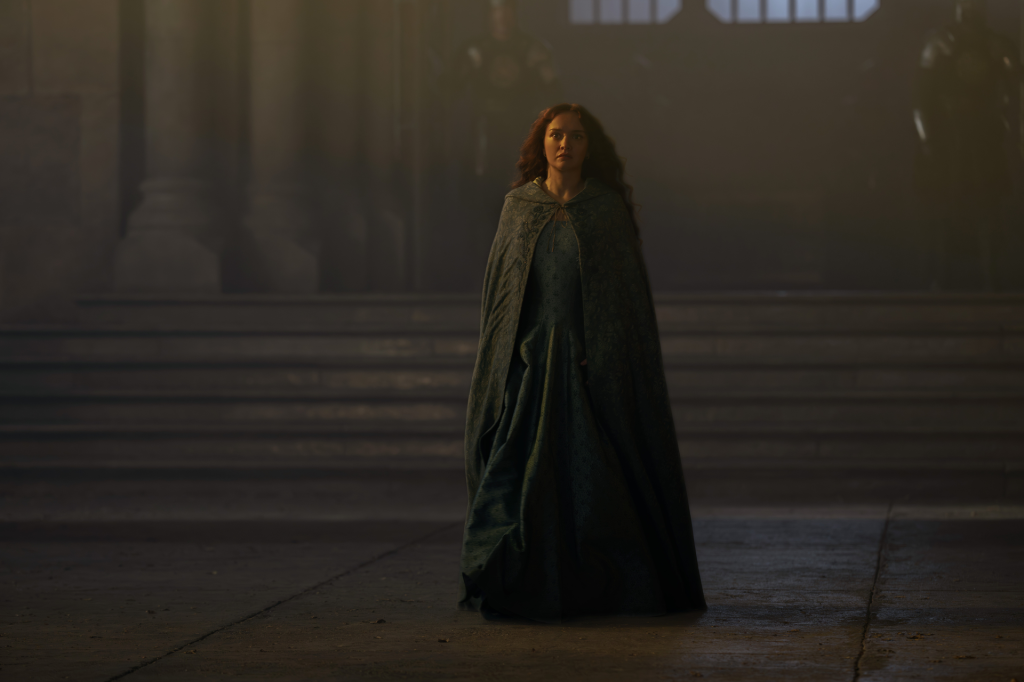 A woman with red hair, wearing a green dress and cape, stands in a dimly lit hall.