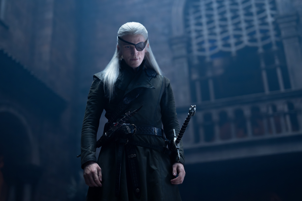 Aemond Targaryen, a man with long white hair and an eye patch, looks directly ahead.