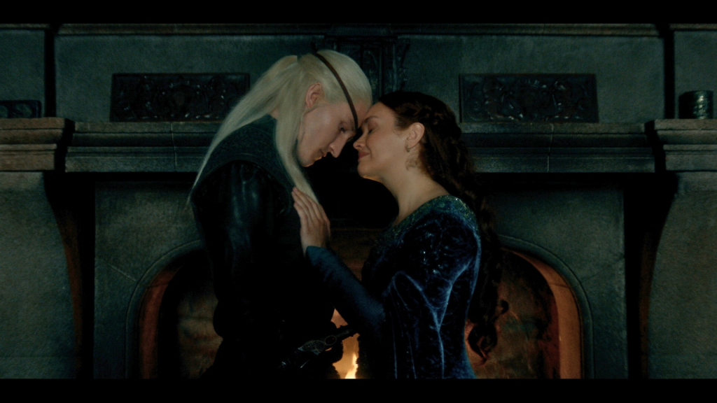 Ewan Mitchell and Olivia Cooke face each other, foreheads touching, in front of a fireplace.