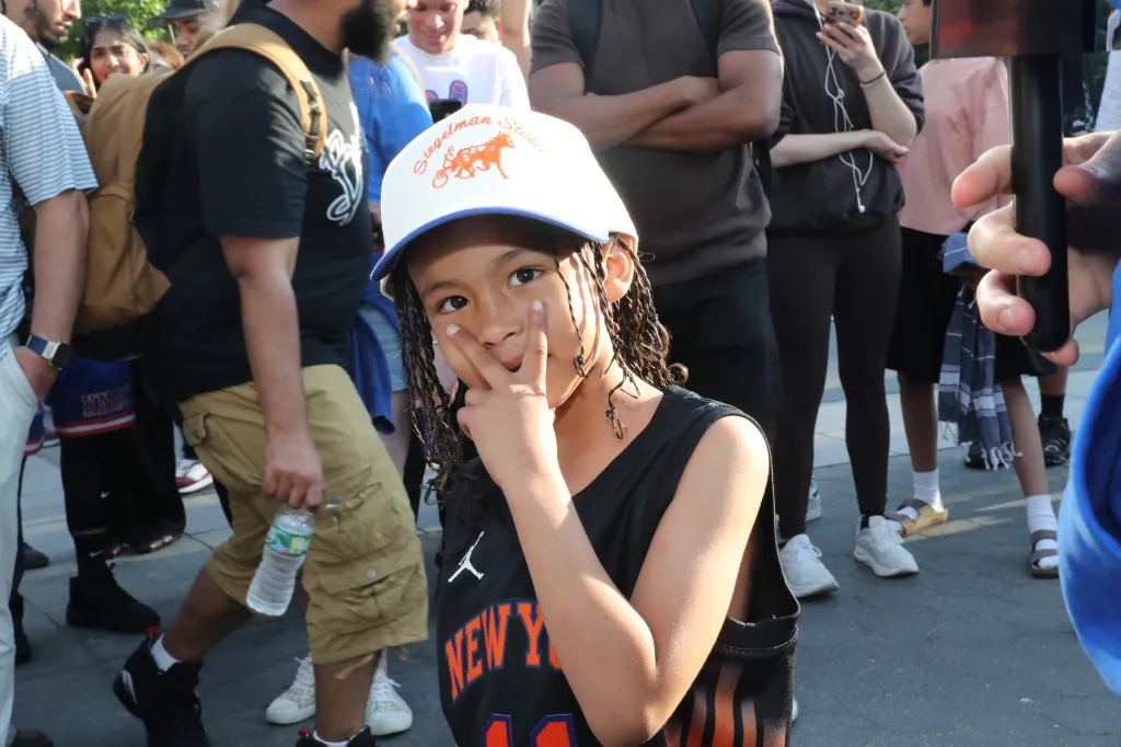 A young boy in a white baseball cap and black New York Knicks jersey making a peace sign with his hand.