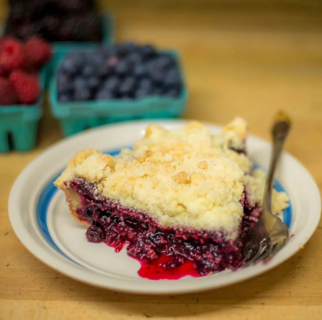 A slice of berry pie with a crumb topping on a plate with a fork, and baskets of fresh berries in the background.