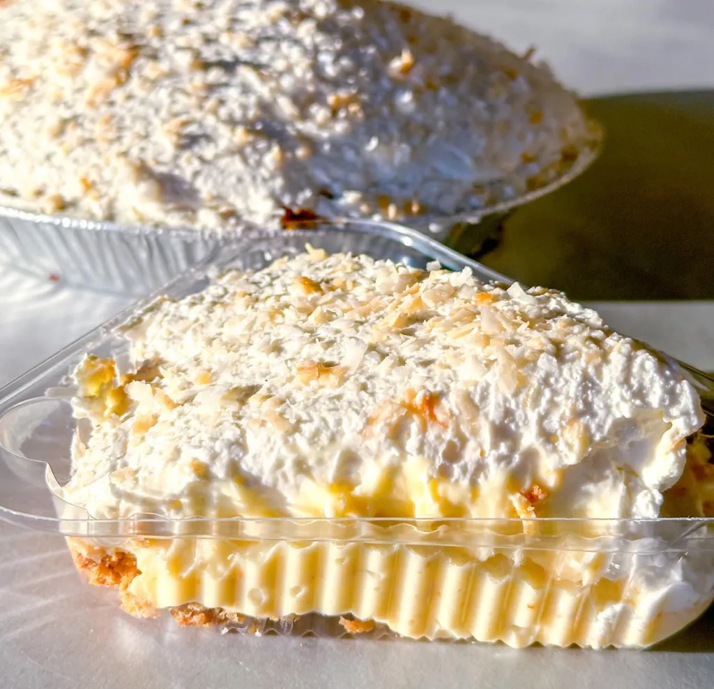 Slice of coconut cream pie in a plastic container with a full pie in the background.