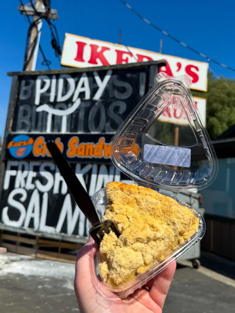 A hand holds a slice of pie in a plastic container with a fork, with an Ikeda's California Country Market sign in the background.
