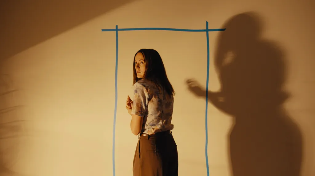 Renate Reinsve looking over her shoulder against a light wall, with a blue rectangle outline and her shadow to the right.