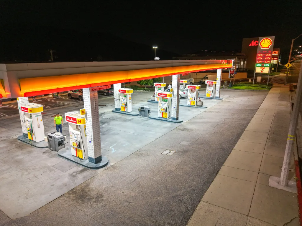 An aerial view of a Shell gas station at night, showing several fuel pumps, high gas prices listed on a large sign, and a lone person near the pumps.