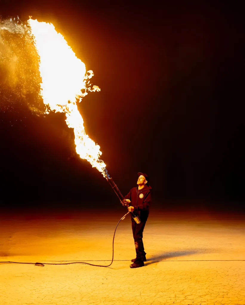 Man in dark clothing and a cap holding a flamethrower, directing a large stream of fire upwards against a dark background.