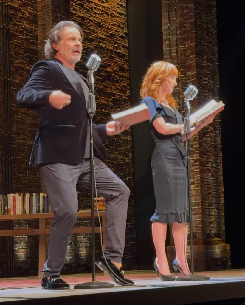 Two actors performing on a stage with microphones and books in hand, with a brick wall backdrop.