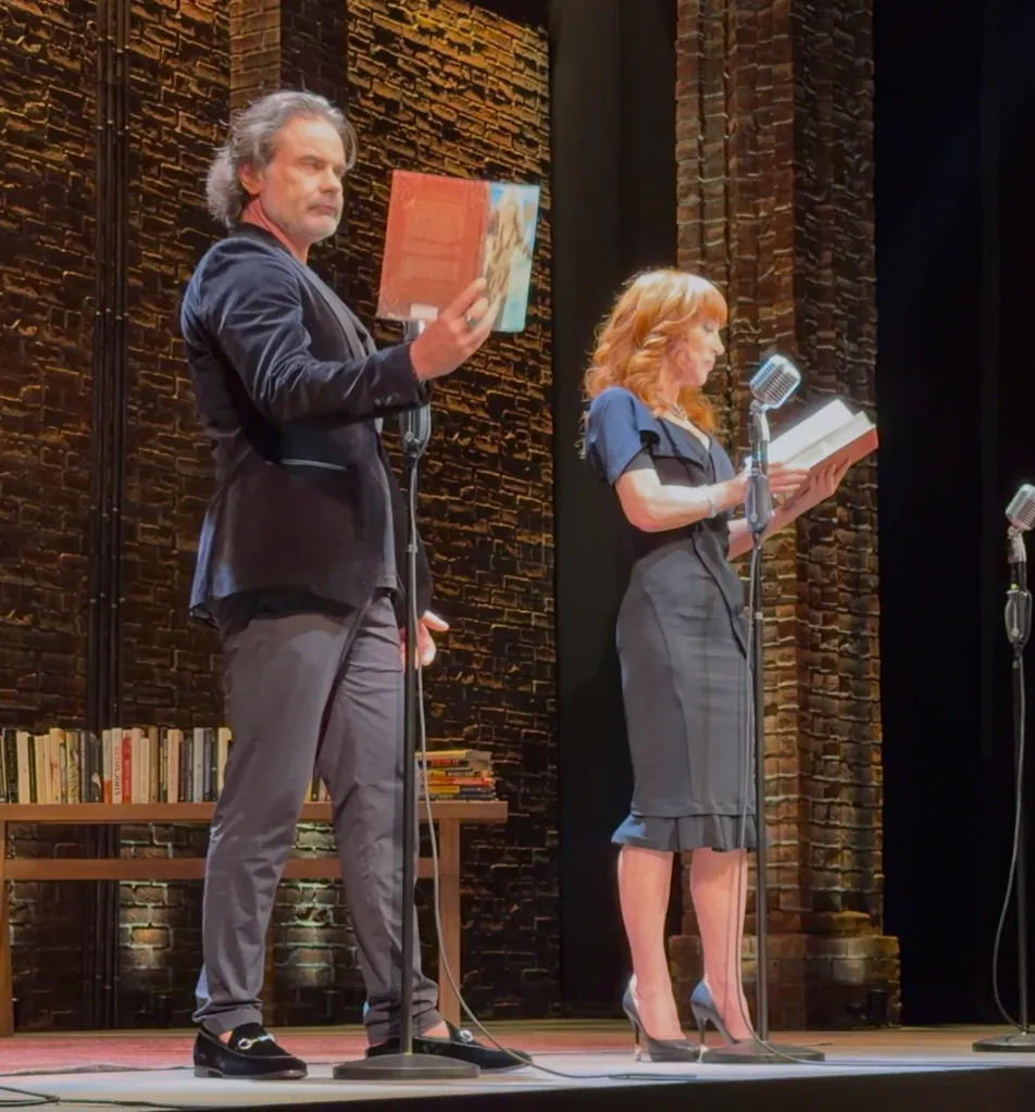 Two people reading books on stage with microphones.