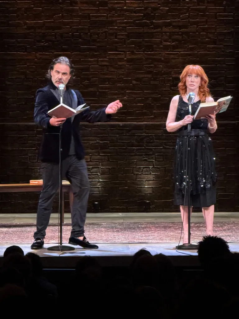 A man and woman standing on a stage, each holding a book and speaking into a microphone.