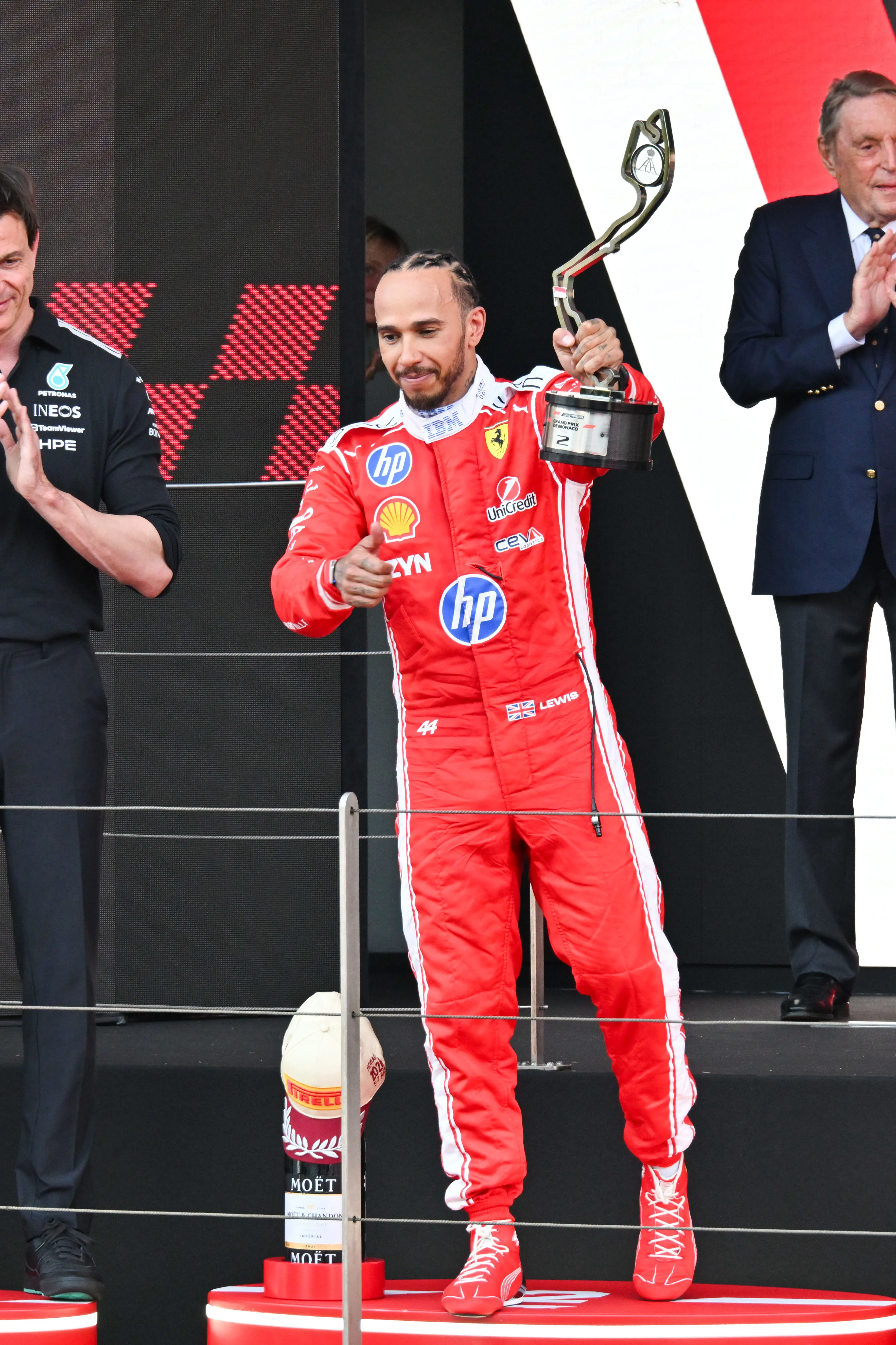 Lewis Hamilton, in a red racing suit, stands on a podium holding a trophy and gesturing to the crowd.
