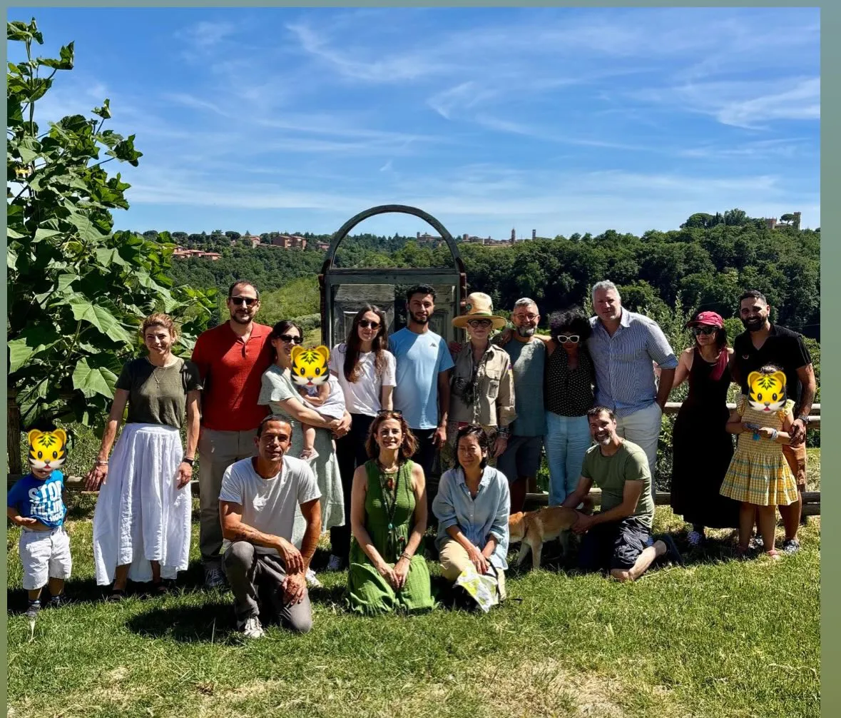 A diverse group of adults and children posing outdoors in a grassy area with trees and buildings in the background.