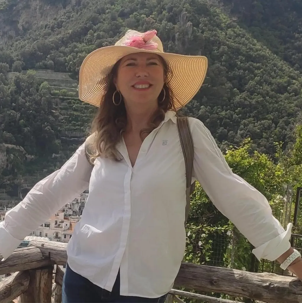 Cinzia Dal Pino wearing a straw hat with pink flowers, smiling against a mountainous backdrop.