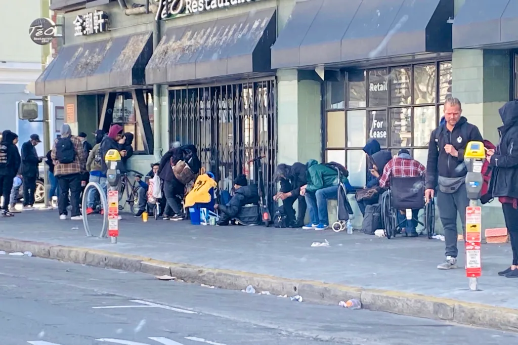 A group of people, some appearing unhoused, gathered on a city sidewalk in front of storefronts.