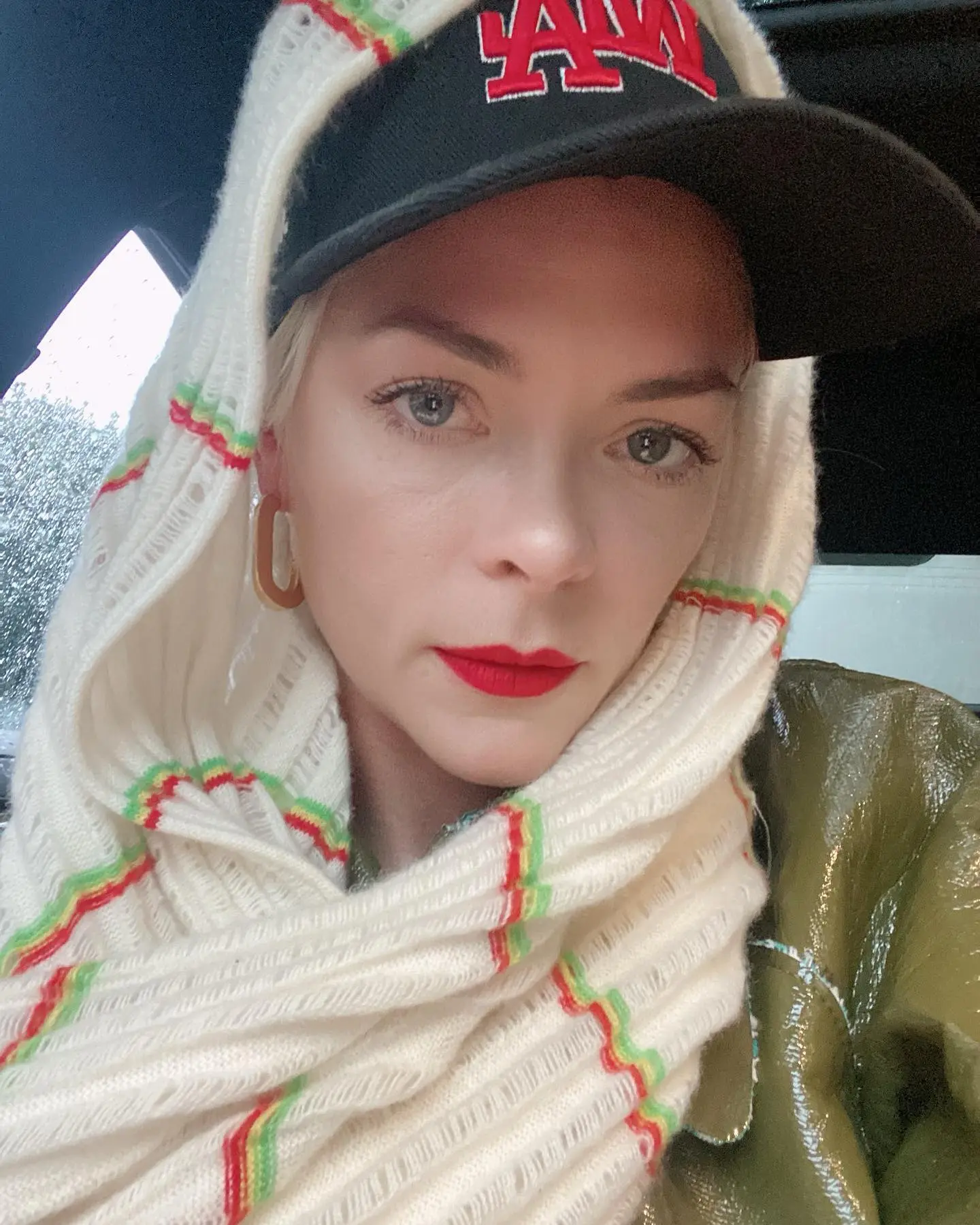 Jaime King in a knitted cream hood with red and green stripes, red lipstick, and a black baseball cap.