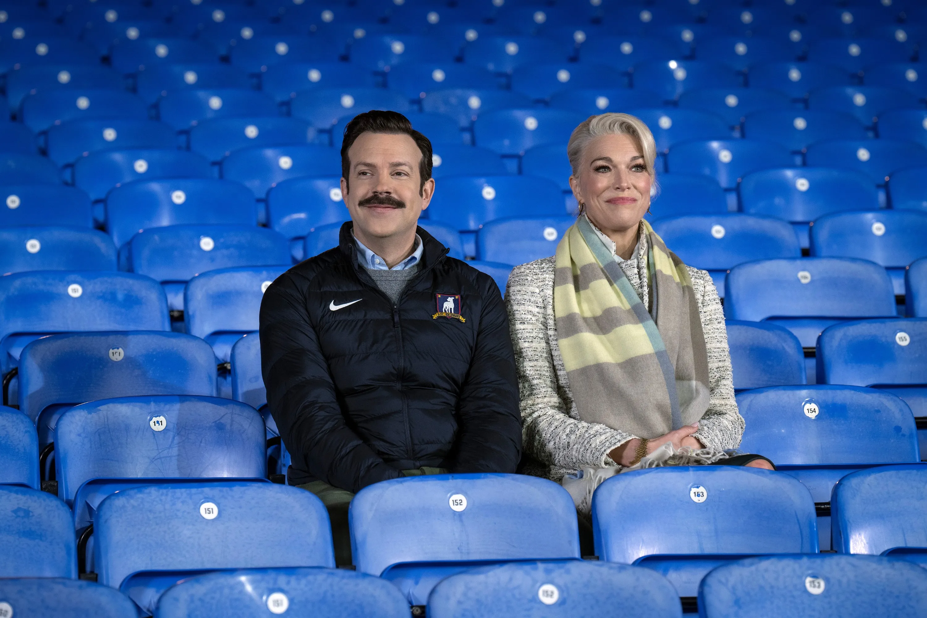 Jason Sudeikis and Hannah Waddingham sitting in blue stadium seats.