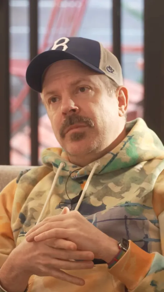 Jason Sudeikis in a blue and white baseball cap and tie-dye hoodie, with his hands clasped together.