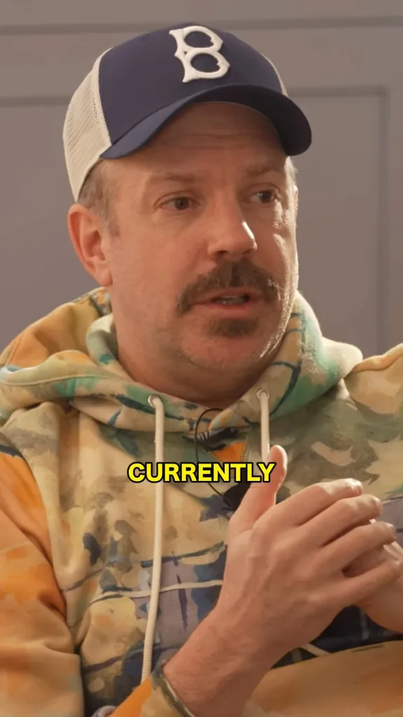 Jason Sudeikis in a blue and white baseball cap and tie-dye hoodie, with the word 