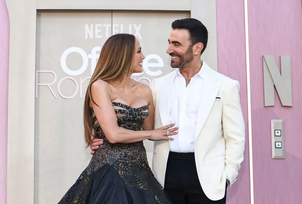 Jennifer Lopez in a black dress and Brett Goldstein in a white suit smile at each other at the 