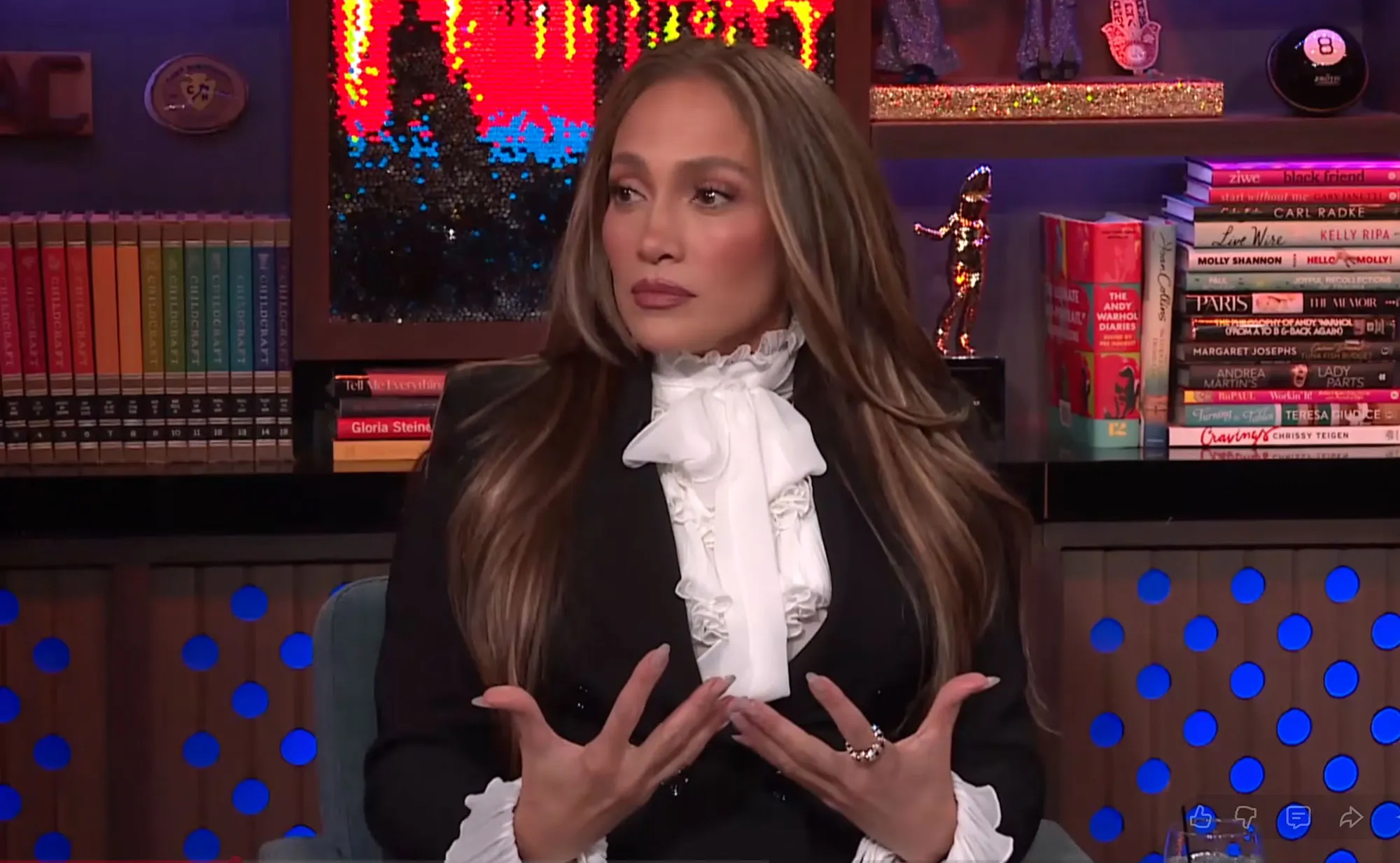 Jennifer Lopez wearing a ruffled white shirt under a black blazer, speaking with her hands open.