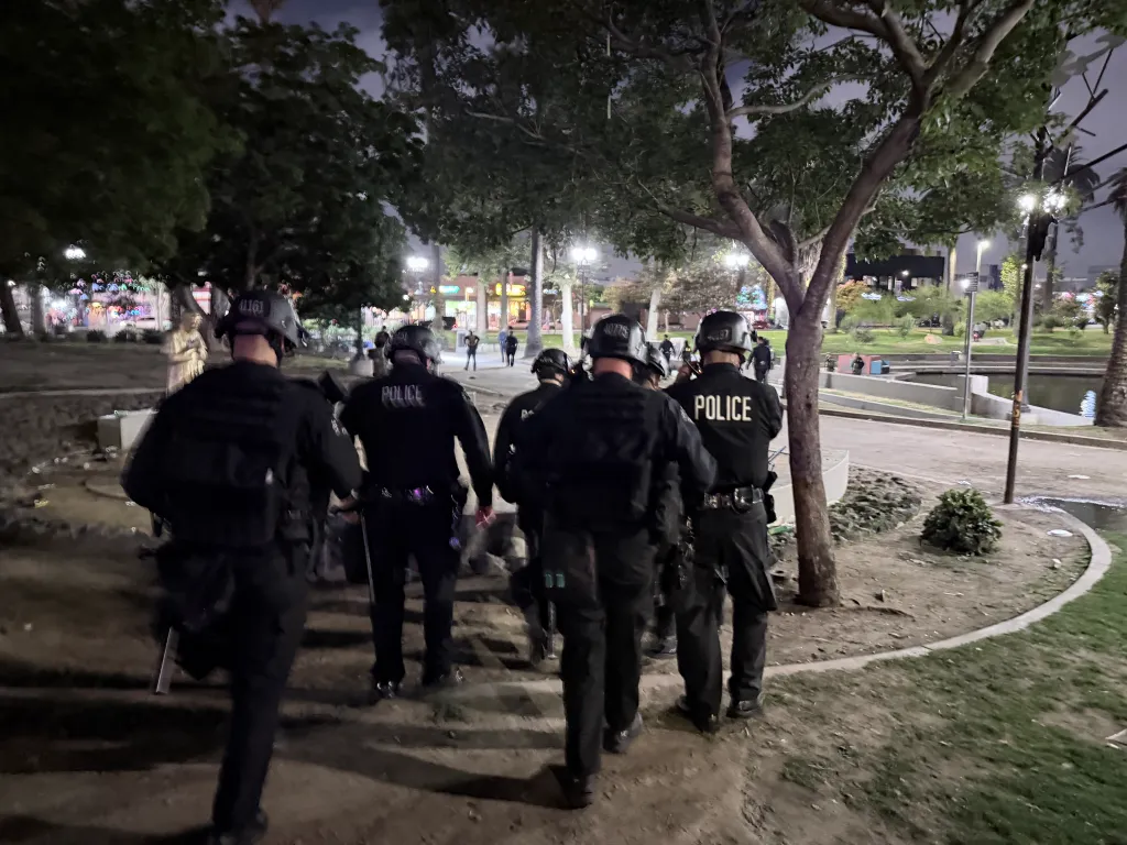 Police officers in tactical gear walk at night through a park littered with debris.