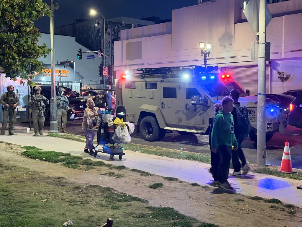 A tactical vehicle with flashing red and blue lights is on a city street at night, with armed police officers standing nearby and people walking past.