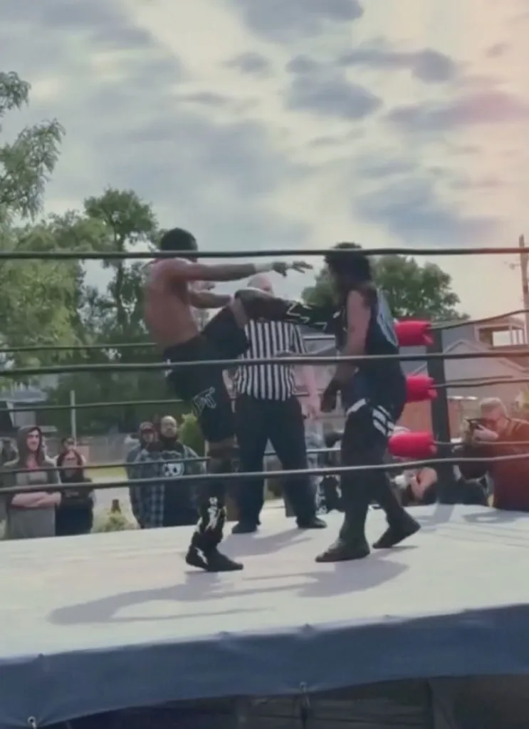Pro wrestler Jordan Saint kicking another wrestler in the ring, with a referee and audience watching.