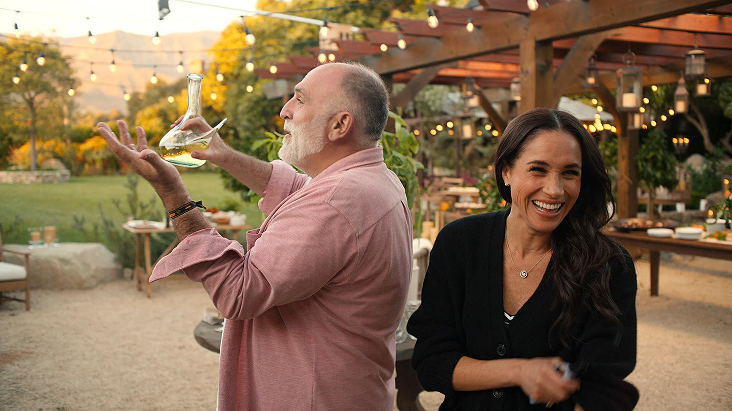 Meghan Markle smiling while Chef José Andrés holds a glass carafe of liquid.