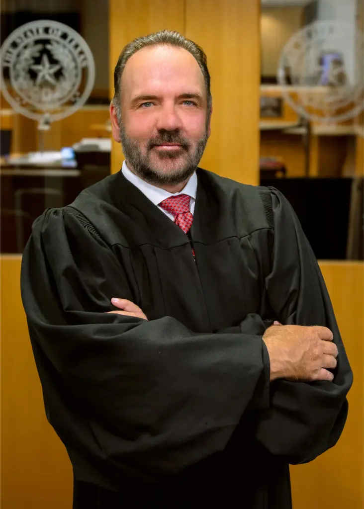 Judge John Roach Jr., wearing a black judicial robe and red tie, stands with arms crossed.