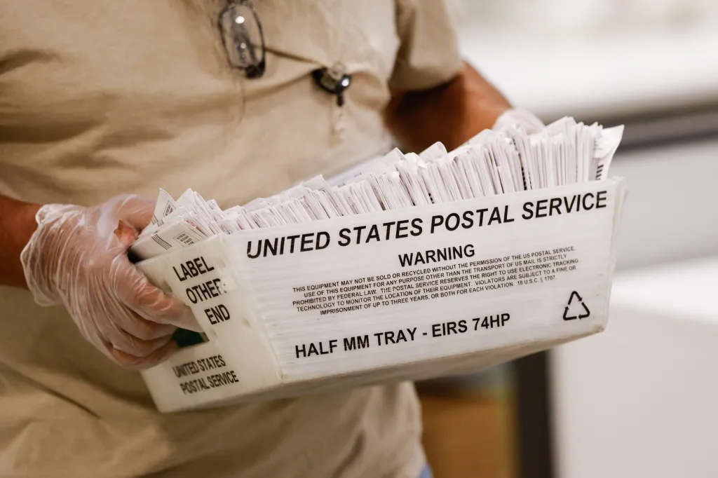 A person wearing gloves carrying a United States Postal Service tray full of ballots.