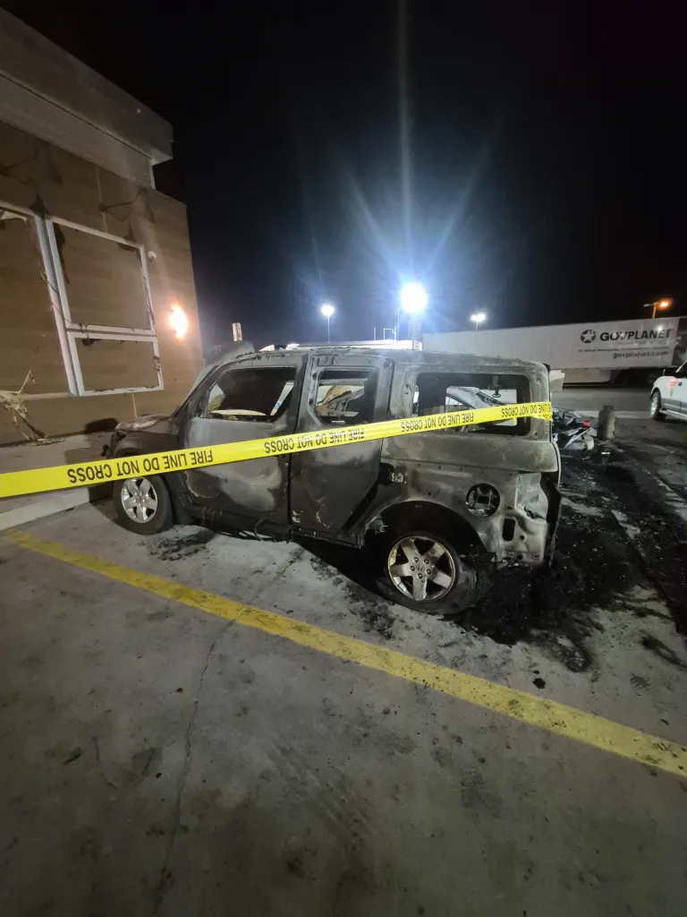 A burnt-out SUV with a shattered windshield, surrounded by fire line tape in a parking lot.