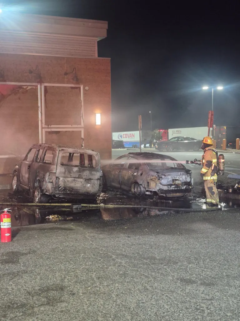 Firefighter hoses down two burnt-out cars in a parking lot at night.