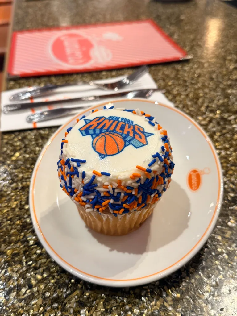A cupcake from Junior's Restaurant and Bakery, topped with white frosting, blue and orange sprinkles, and the New York Knicks logo.