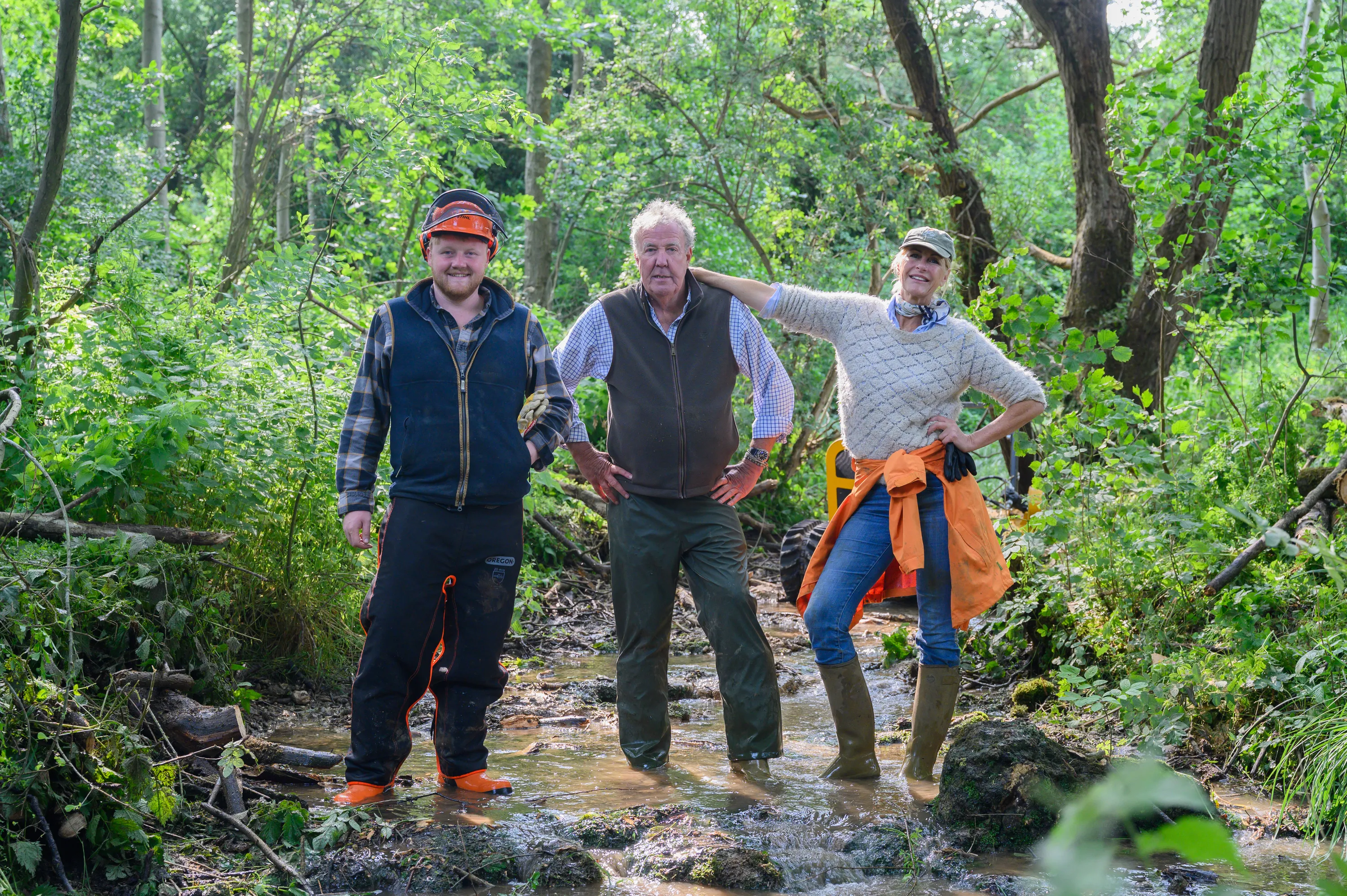 Kaleb Cooper, Jeremy Clarkson, and Lisa Hogan standing in a muddy stream within a forest.