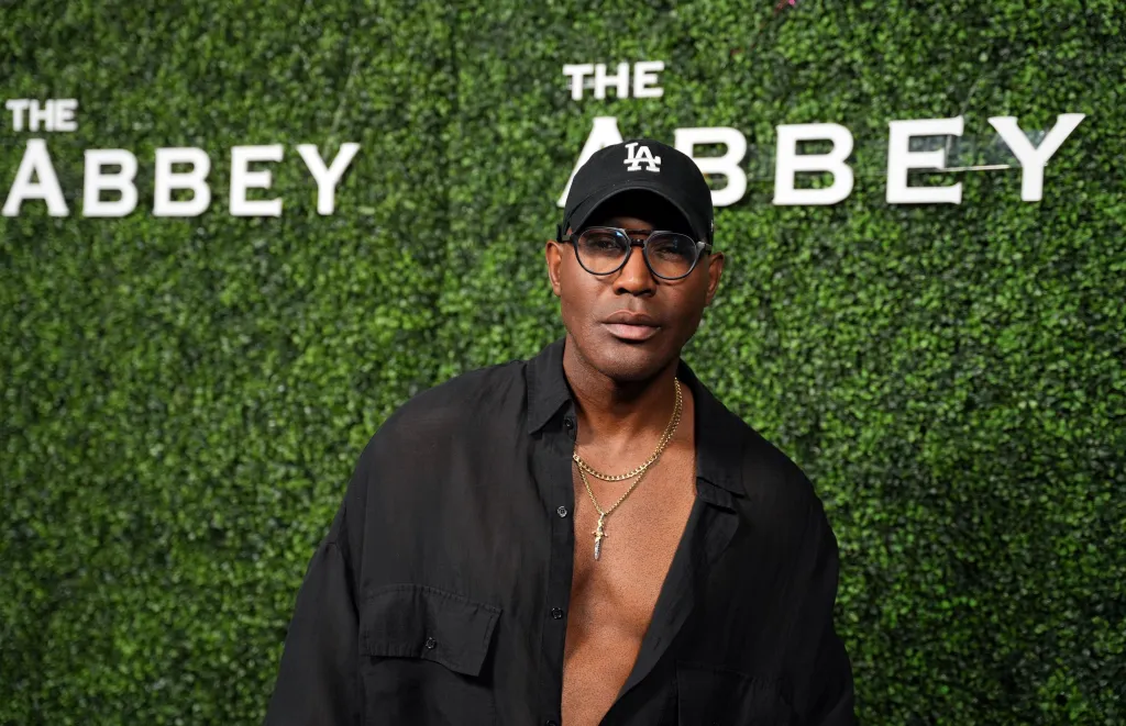 Karamo Brown wearing a black cap, glasses, and an open black shirt with gold chains against a green hedge wall with
