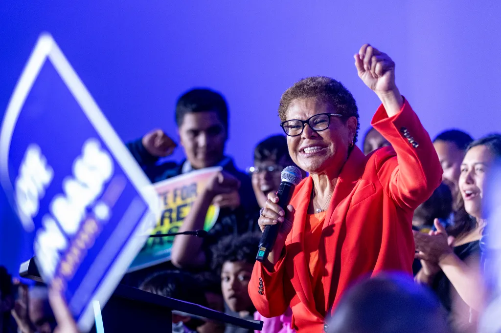 Karen Bass smiling and raising her fist while holding a microphone at an election night party.